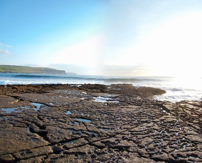 Rocky limestone coastal platform with water-filled crevices extending to ocean waves, with green cliffs visible on the horizon under bright sun