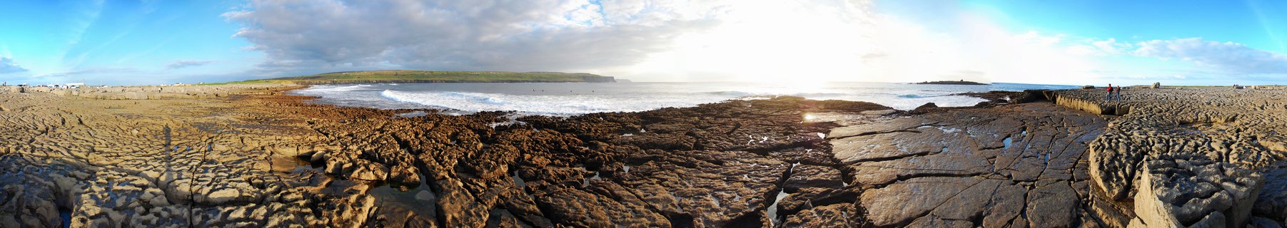 Wide coastal landscape showing cracked limestone pavement in foreground with ocean waves and distant green hills under cloudy sky with sun breaking through