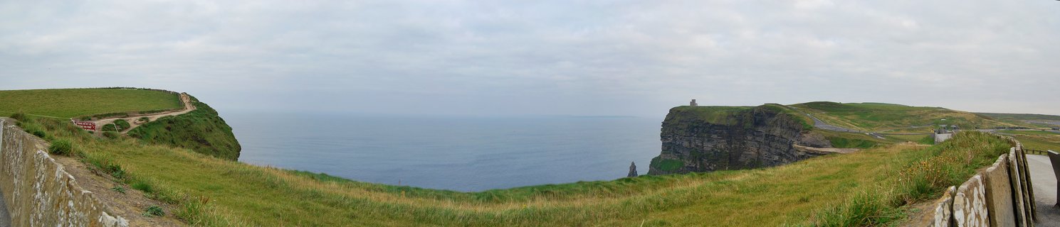 Panoramic view of Cliffs of Moher showing green grassy clifftops, stone walls, O'Brien Tower on distant cliff, and Atlantic Ocean under overcast sky