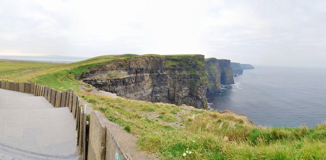 Coastal cliffs with layered rock faces covered in green grass overlooking the Atlantic Ocean, with wooden safety fencing and viewing platform visible in foreground