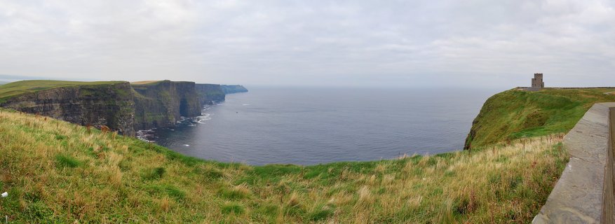 Panoramic view of Cliffs of Moher showing steep rock faces meeting the Atlantic Ocean, with green grassy slopes in the foreground and O'Brien's Tower on the right cliff edge under cloudy sky