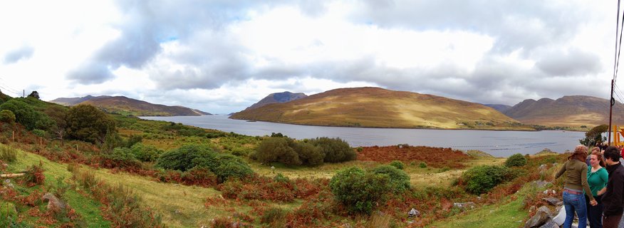 Panoramic landscape showing a lake surrounded by brown and green hills in Connemara, Ireland, with three people standing at a viewpoint on the right side under cloudy skies