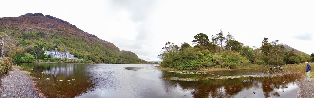 A panoramic view of Kylemore Abbey, a white castle-like building on the shore of a lake with mountains covered in green vegetation rising behind it in Connemara, Ireland