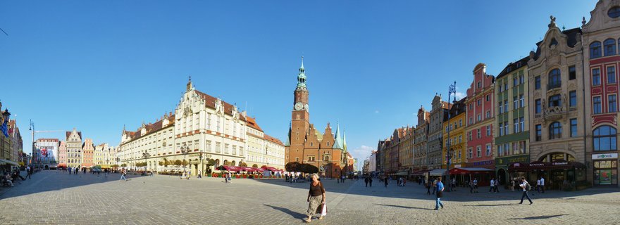 Wide panoramic view of Wrocław Market Square showing the Gothic brick Town Hall clock tower centered among colorful historic buildings with pedestrians walking across cobblestone plaza under clear blue sky