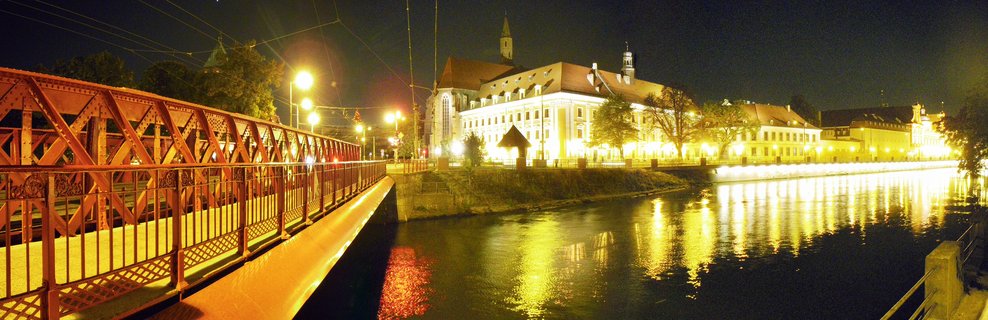 Night panorama of Wrocław showing a red metal bridge over a river with illuminated historic buildings and church towers reflecting in the water