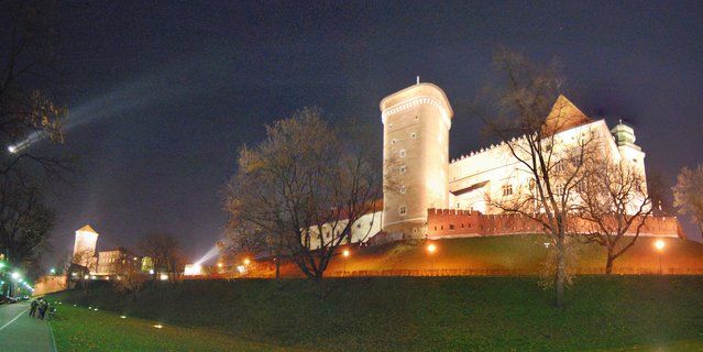 Night photograph of Wawel Castle in Krakow showing illuminated medieval stone towers and fortified walls on a grassy hill with bare trees in the foreground