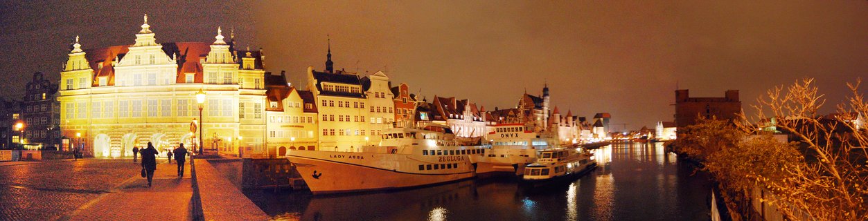 Night photograph of Gdańsk's historic waterfront showing illuminated multi-story buildings with ornate gables along a river, several white boats docked at the quay, people walking on a stone promenade, warm yellow lighting reflecting in dark water