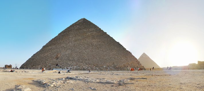 The Great Pyramid of Giza rises from desert sand with tourists and camels at its base under a clear blue sky