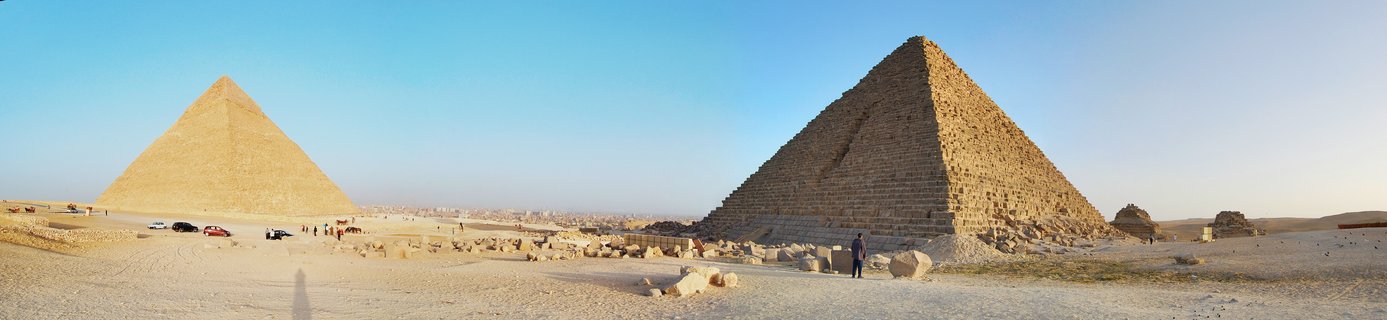 Two large ancient Egyptian pyramids on desert sand under clear blue sky with tourists and vehicles in foreground