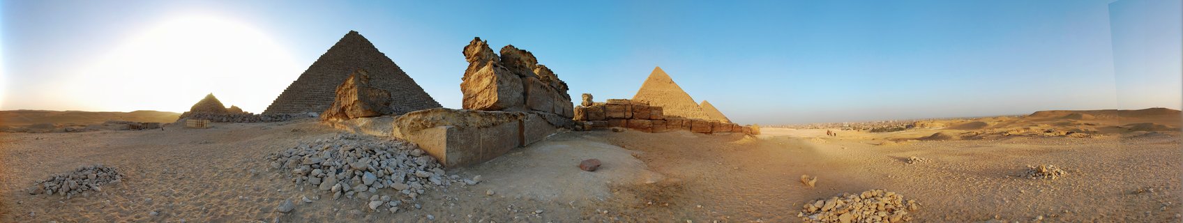 Panoramic photograph of the Giza Pyramids in Egypt showing three major pyramids and ancient stone ruins in desert landscape during golden hour with clear blue sky