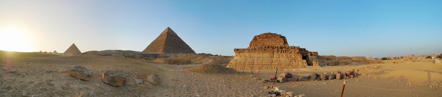 Panoramic view of the three Giza pyramids at sunset with ancient stone ruins in the foreground on desert sand under clear blue sky
