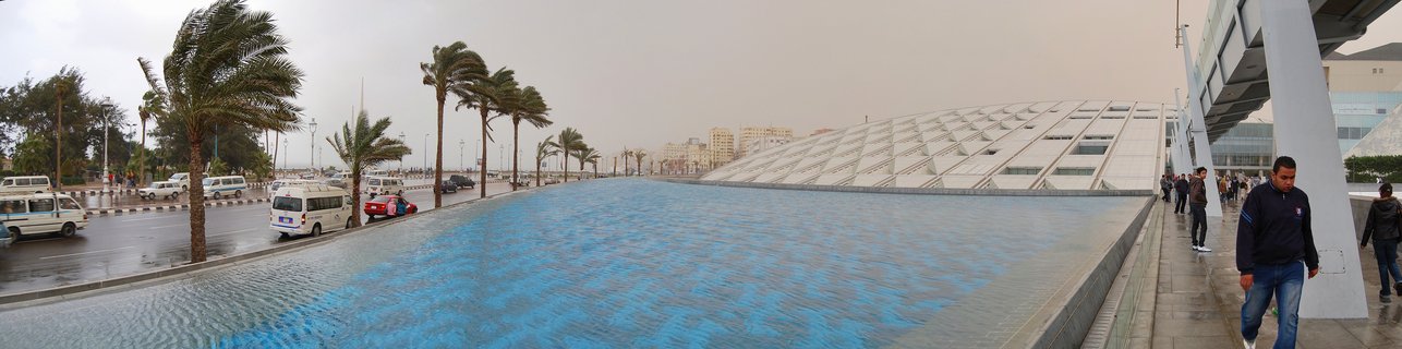 Wide view of the Bibliotheca Alexandrina library building with distinctive slanted geometric roof, large blue reflecting pool in foreground, palm trees bending in wind, vehicles on adjacent road, pedestrians walking on sidewalk under overcast sky