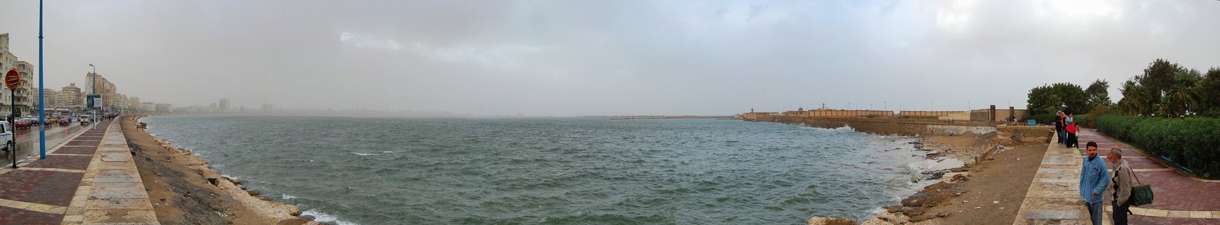 Panoramic view of Alexandria's waterfront corniche showing the Mediterranean Sea, stone seawall, promenade with pedestrians, coastal buildings, and overcast sky