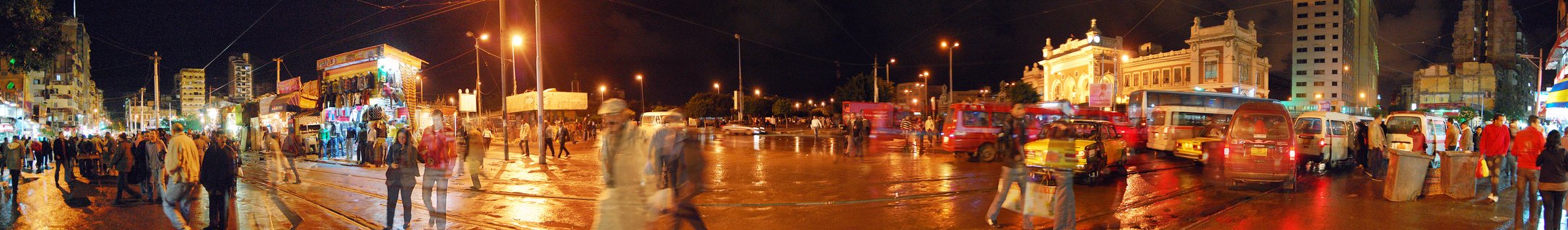 Nighttime panoramic street view of Alexandria, Egypt showing pedestrians walking on wet pavement, multiple buses and vehicles, illuminated buildings with colonial architecture, and streetlights reflecting on the road surface