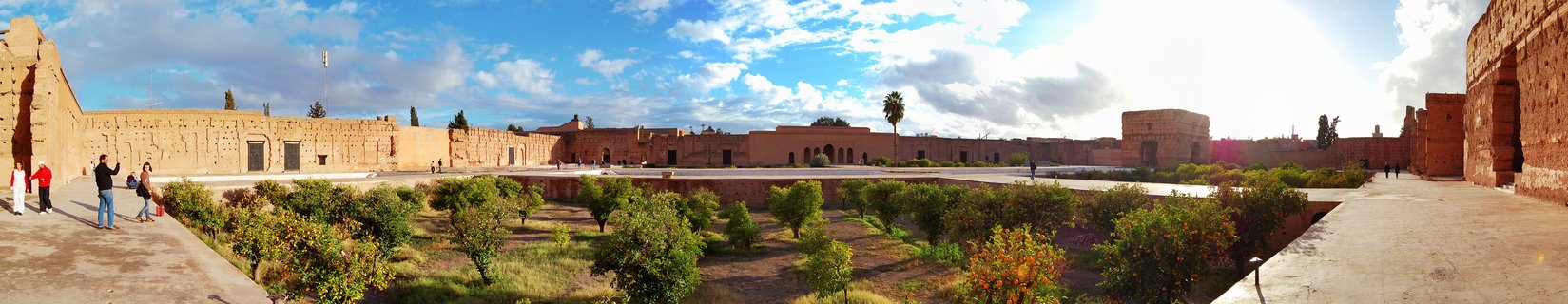 Panoramic view of El Badi Palace ruins in Marrakesh showing red clay walls surrounding a sunken garden with small trees, tourists walking on stone pathways under blue sky with clouds