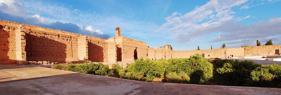 Wide view of ancient reddish-brown earthen palace walls with rectangular openings, green bushes in foreground, blue sky with white clouds above, Marrakesh Morocco