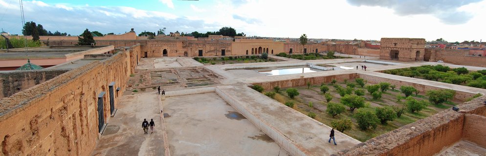 Aerial view of El Badi Palace courtyard in Marrakesh showing weathered ochre walls, sunken gardens with rows of orange trees, reflection pools, and visitors walking on stone pathways under cloudy sky