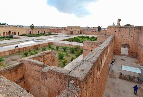 Aerial view of El Badi Palace ruins showing reddish-brown stone walls surrounding sunken gardens with green citrus trees and rectangular reflecting pools, with visitors walking on the grounds