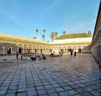 Wide courtyard of Bahia Palace in Marrakesh with traditional Moroccan architecture, green-tiled roofs, arched colonnades, visitors walking on geometric tile pavement, palm trees, and blue sky