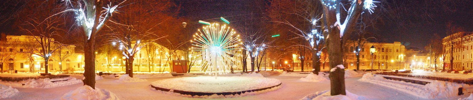 Night photograph of a snow-covered park with an illuminated fountain featuring green and white starburst lights, surrounded by trees decorated with white string lights and historic buildings in the background
