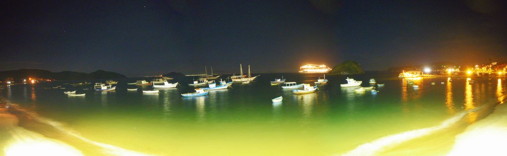 Night photograph of a harbor with numerous fishing boats anchored in calm water, illuminated buildings along the shoreline, and dark hills in the background under a night sky