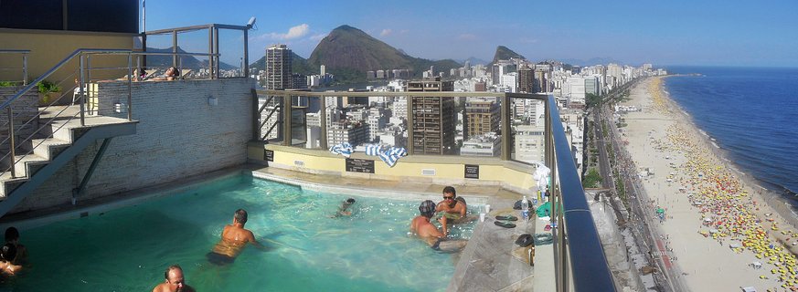 People swimming in a rooftop pool with a panoramic view of Copacabana Beach, high-rise buildings, and Sugarloaf Mountain in Rio de Janeiro, Brazil.