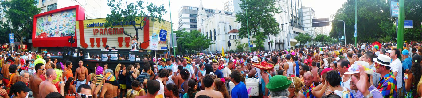 Large crowd of people in colorful costumes and hats celebrating at a Carnival street party in Rio de Janeiro with a red and yellow sound truck displaying Ipanema