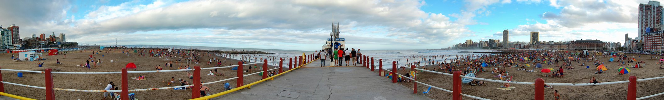 Wide panoramic photograph of a crowded beach in Mar del Plata, Argentina, showing a central pier walkway with red posts and white railings, hundreds of beachgoers with colorful umbrellas on sand, ocean waves, and city buildings along the coastline under a partly cloudy sky
