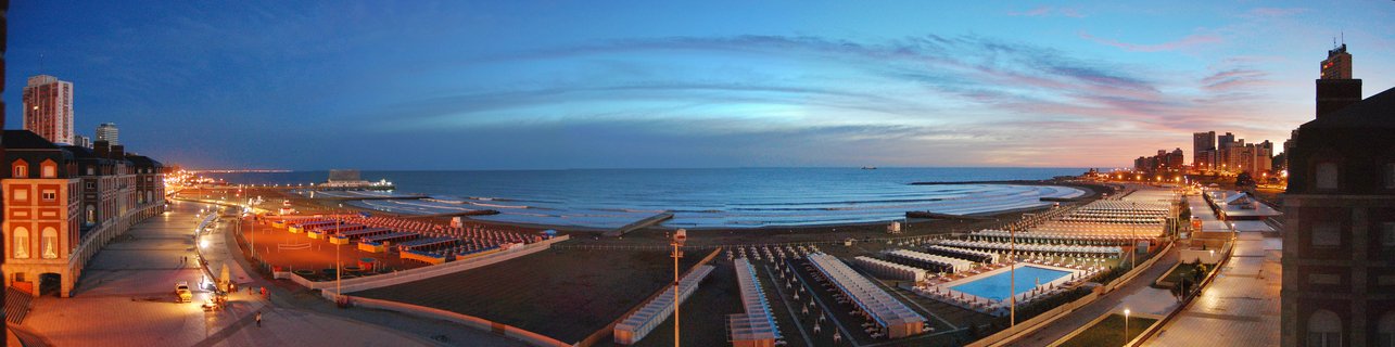 Panoramic view of Mar del Plata coastline at blue hour showing organized rows of beach umbrellas and cabanas, illuminated swimming pool complex, Atlantic Ocean waves, glowing street lights along promenade, and hotel buildings against twilight sky in Buenos Aires Province, Argentina