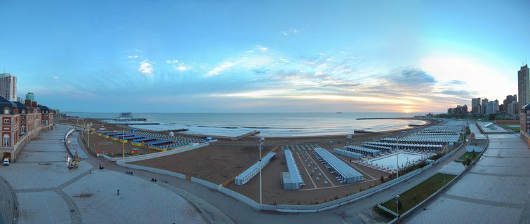 Panoramic aerial view of Mar del Plata beach at sunrise showing organized white beach cabanas, curved promenade, ocean horizon, and coastal buildings under blue sky with scattered clouds