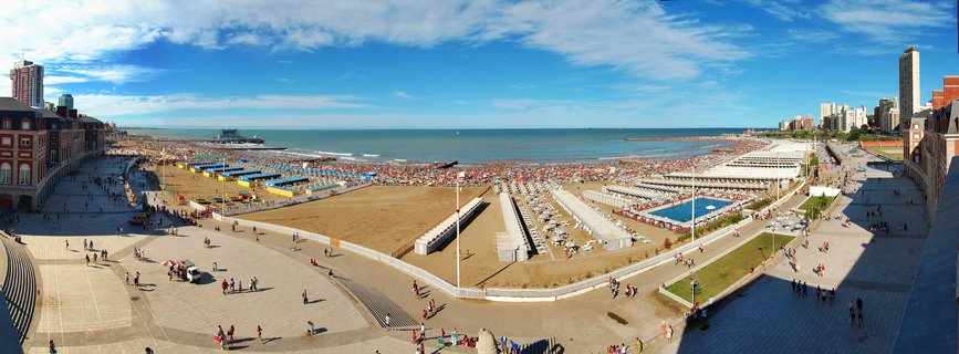 Panoramic aerial photograph of a crowded beach in Mar del Plata, Argentina, showing colorful umbrellas on sand, Atlantic Ocean, blue sky with clouds, paved walkways with people, and buildings along the coastline