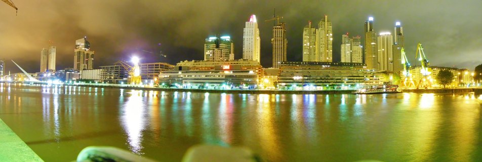 Night cityscape showing illuminated skyscrapers along a waterfront with reflections in calm water and yellow port cranes on the right side