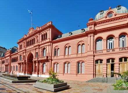 Pink neoclassical government building with Argentine flag, fountains and planters in foreground, clear blue sky