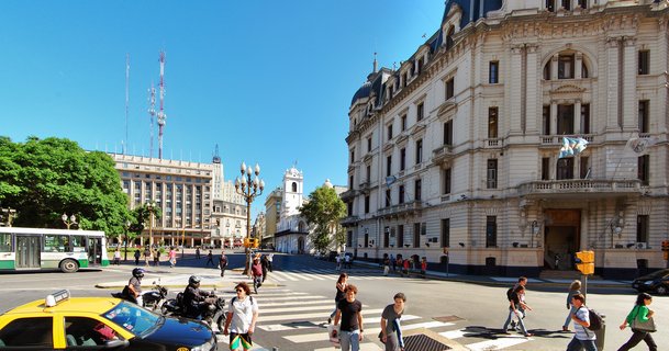 Street intersection in Buenos Aires showing pedestrians crossing, yellow taxi, city bus, historic buildings with Argentine flags, and a white church tower under blue sky