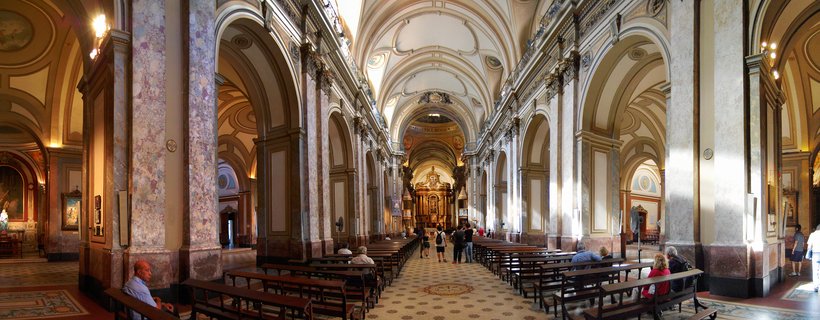 Wide interior view of a cathedral showing rows of marble columns, wooden pews, vaulted ceiling with decorative elements, and people walking toward an ornate altar at the far end