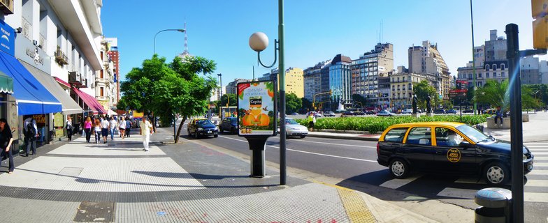 Wide street scene in Buenos Aires showing a black and yellow taxi, pedestrians walking on sidewalks, street lamps, advertising kiosk, trees, and multiple buildings under a clear blue sky