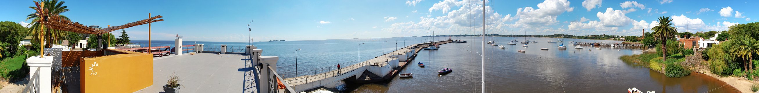 Panoramic waterfront view of Colonia del Sacramento harbor in Uruguay showing boats in marina, pier, terrace with orange wall, palm trees, and Rio de la Plata under blue sky with clouds