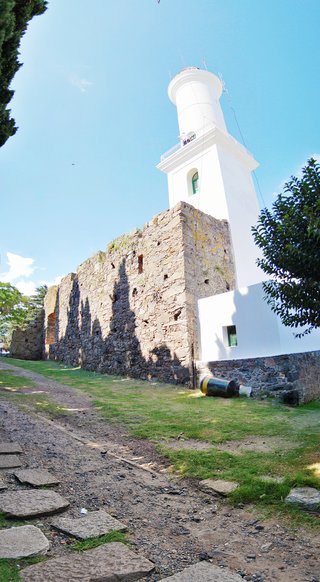 White cylindrical lighthouse tower rising from historic stone fortress walls with grassy foreground and clear blue sky in Colonia del Sacramento