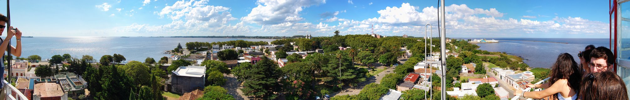 Wide panoramic aerial photograph showing Colonia del Sacramento coastal town in Uruguay with colonial buildings, green trees, blue water of Rio de la Plata, and cloudy sky