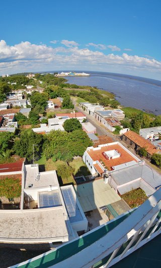 Aerial photograph showing colonial buildings with red tile roofs and white walls in Colonia del Sacramento, Uruguay, with green trees, cobblestone streets, the Río de la Plata river, and a large passenger ferry visible in the distance under blue sky with scattered clouds.