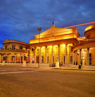 Teatro Solís theater building in Montevideo at night with golden illumination, columns, blue sky, and red light trails from traffic on cobblestone street