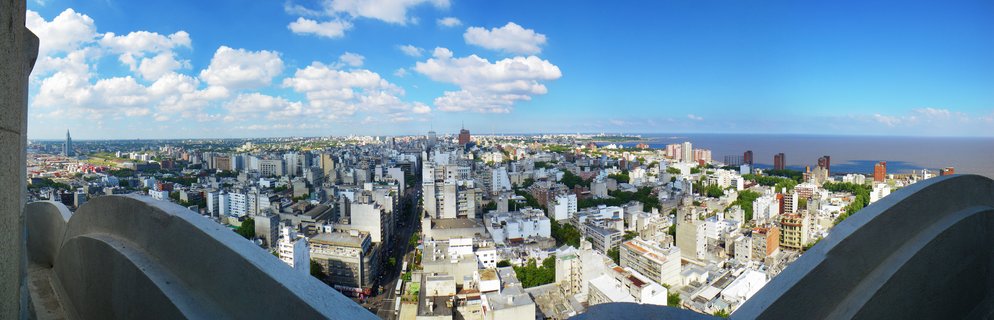 Panoramic cityscape of Montevideo, Uruguay showing dense urban buildings stretching to the Rio de la Plata coastline under blue sky with white clouds, viewed from an elevated stone structure