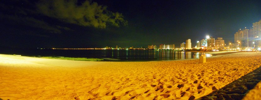 Night photograph of a beach with golden-lit sand in the foreground, dark ocean water with city light reflections, and illuminated high-rise buildings along the coastline under a dark sky with clouds