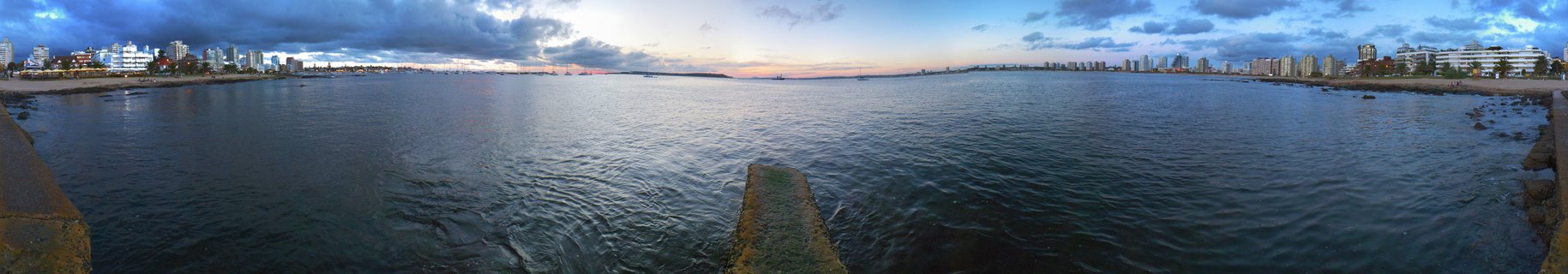 Wide panoramic photograph of Punta del Este bay at dusk showing city buildings along the coastline under a dramatic cloudy sky with water in the foreground