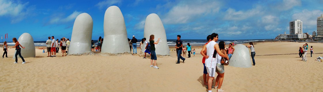 Large white finger-shaped sculptures emerging from sandy beach with tourists walking around and ocean waves in background under blue sky