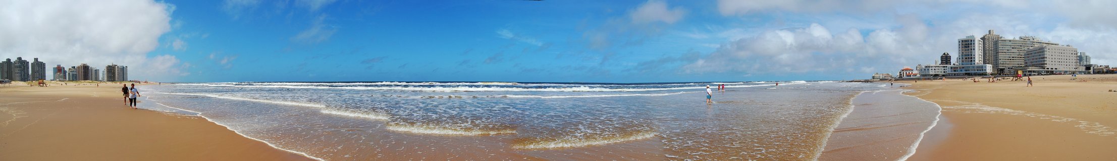 Wide beach panorama showing sand, ocean waves, scattered beachgoers, and high-rise buildings along the coastline under blue sky with clouds