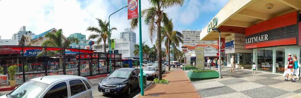 Wide street view showing palm trees, parked cars, pedestrians walking past storefronts including a real estate office and Burger King sign, with multi-story buildings in the background under a partly cloudy blue sky