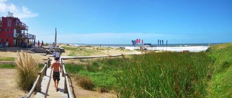 A shirtless man walks on a wooden boardwalk toward a beach with waves, a red multi-story building on the left, green beach grass in the foreground, and blue sky above at Punta del Diablo, Uruguay