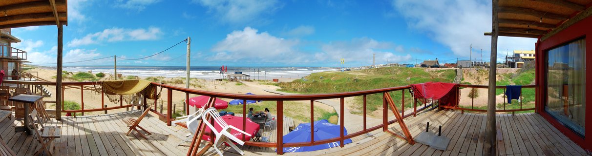 Wooden deck terrace with colorful umbrellas overlooking sandy beach and ocean waves under blue sky with clouds in Punta del Diablo, Uruguay