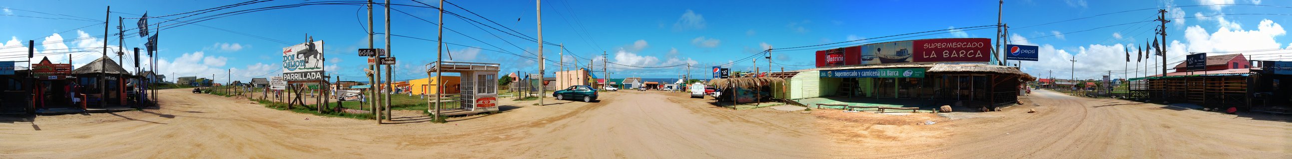 Panoramic street scene showing dirt roads, small shops including Supermercado La Barca, utility poles, and buildings in Punta del Diablo, Uruguay under blue sky with clouds